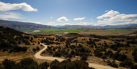 Backcountry nature panorama of road winding through dry New Zealand landscape and green agriculture fields in background