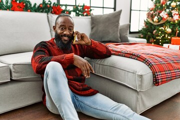 Obraz premium Young african american man talking on the smartphone sitting by christmas tree at home