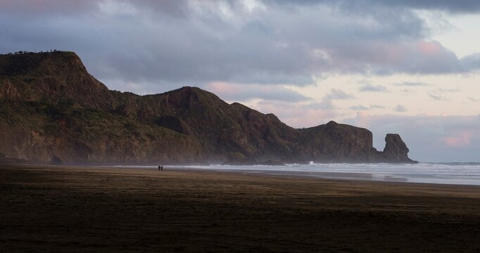 A couple walking along idyllic black sand Te Henga Bethells beach with rock formation during sunset in New Zealand