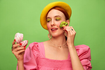 Beautiful young girl in cute outfit with yellow beret eating broccoli and meringue on green background. Food pop art photography. Complementary colors. Concept of art, beauty, food. Copy space for ad