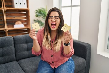 Fototapeta premium Young hispanic woman holding sad to happy emotion paper angry and mad screaming frustrated and furious, shouting with anger. rage and aggressive concept.