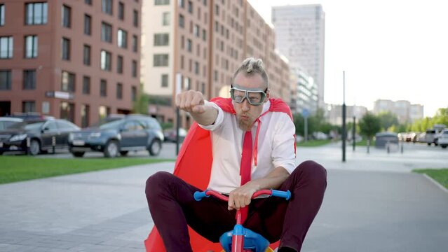 Portrait of young businessman in suit wearing superman outdoors on bicycle. Leader success winner superhero crazy man with red cape riding bicycle on city street, business. People and work concept.