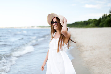 Fototapeta na wymiar Happy smiling woman in free happiness bliss on ocean beach standing and posing with hat and sunglasses. Portrait of a female model in white summer dress enjoying nature 
