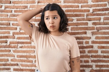 Young hispanic woman standing over bricks wall confuse and wondering about question. uncertain with...