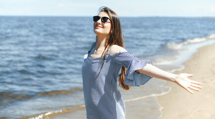 Happy smiling woman in free bliss on ocean beach standing with open hands. Portrait of a brunette female model in summer dress enjoying nature during travel holidays vacation outdoors