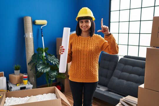 Young hispanic woman standing on new home with blueprints surprised with an idea or question pointing finger with happy face, number one