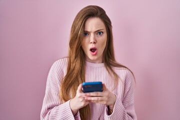 Young caucasian woman using smartphone typing message in shock face, looking skeptical and...
