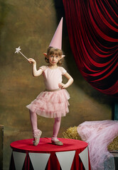 Naklejka premium Magic. Little cute girl, kid in image of magical elf posing in pink dress over dark circus backstage background. Retro circus performance. Concept of dreams, art, fashion, vintage style.