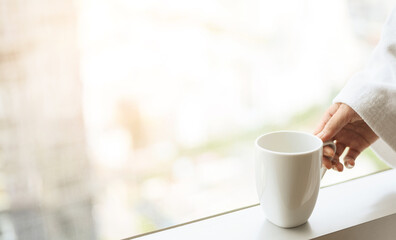 Close up to woman hand in bathrobe holding white cup of coffee near the window, enjoy wonderful morning in the hotel, detaching from work.