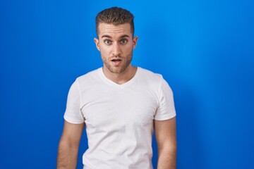Obraz premium Young caucasian man standing over blue background in shock face, looking skeptical and sarcastic, surprised with open mouth