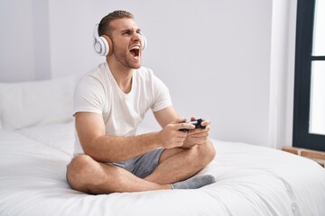 Young caucasian man sitting on the bed at home playing video games angry and mad screaming frustrated and furious, shouting with anger looking up. © Krakenimages.com