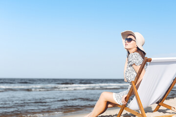 Happy brunette woman wearing sunglasses and hat relaxing on a wooden deck chair at the ocean beach