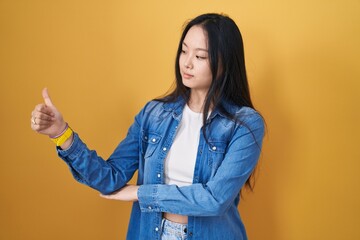 Young asian woman standing over yellow background looking proud, smiling doing thumbs up gesture to the side