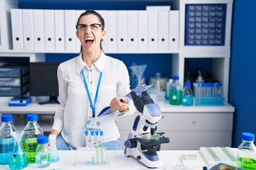 Young brunette woman working at scientist laboratory angry and mad screaming frustrated and furious, shouting with anger. rage and aggressive concept.