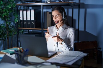 Young brunette woman wearing call center agent headset working late at night pointing fingers to...