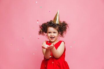 a little girl in a red dress celebrates her birthday, blowing and catching confetti on a pink background.