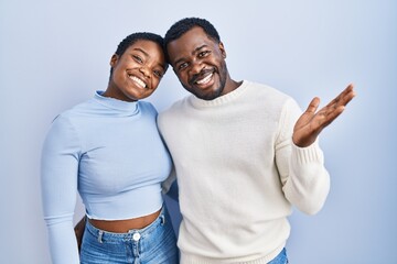 Young african american couple standing over blue background smiling cheerful presenting and pointing with palm of hand looking at the camera.