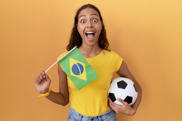 Young hispanic woman holding brazil flag and football ball celebrating crazy and amazed for success with open eyes screaming excited.