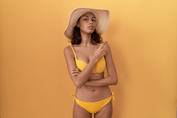 Young hispanic woman wearing bikini and summer hat pointing with hand finger to the side showing advertisement, serious and calm face
