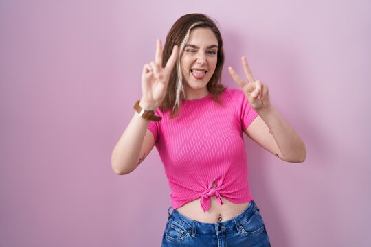Blonde caucasian woman standing over pink background smiling with tongue out showing fingers of both hands doing victory sign. number two.