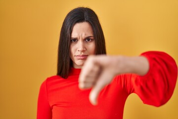 Young hispanic woman standing over yellow background looking unhappy and angry showing rejection and negative with thumbs down gesture. bad expression.