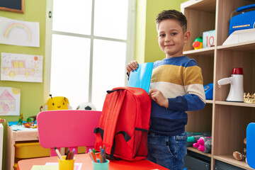 Adorable hispanic boy student holding book of backpack at kindergarten