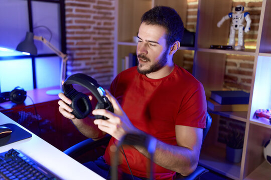 Young hispanic man streamers sitting on table holding headphones at gaming room