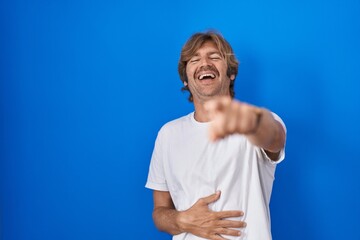 Naklejka premium Middle age man standing over blue background laughing at you, pointing finger to the camera with hand over body, shame expression