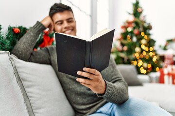 Young hispanic man reading book sitting on sofa by christmas tree at home