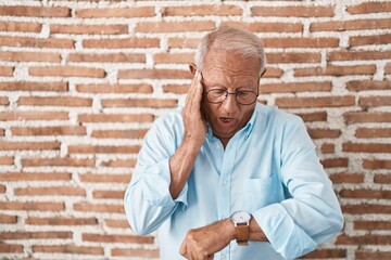 Senior man with grey hair standing over bricks wall looking at the watch time worried, afraid of...