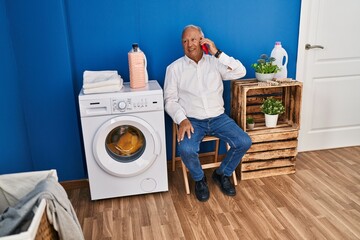 Senior man talking on the smartphone waiting for washing machine at laundry room