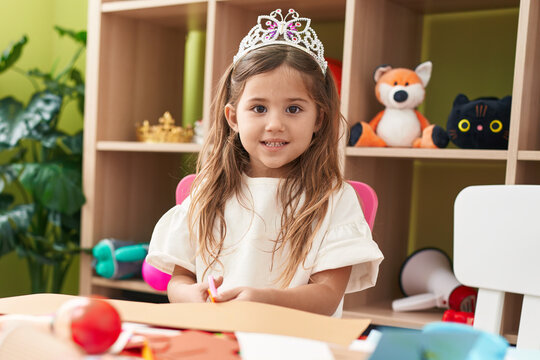 Adorable blonde girl student wearing princess crown cutting paper at kindergarten