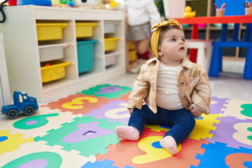 Adorable blonde toddler sitting on floor with serious expression at kindergarten