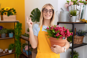 Young caucasian woman working at florist shop holding plant smiling happy pointing with hand and finger to the side