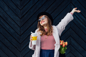 Happy, emotional young fashion woman with reusable coffee cup and net bag with fresh tulip flowers inside on the black wooden background. Urban city street fashion. Spring mood. Selective focus