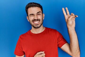Young hispanic man with beard wearing red t shirt over blue background smiling with happy face...