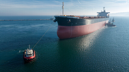 Aerial view of tug boat assisting big cargo ship. Large cargo ship enters the port escorted by...
