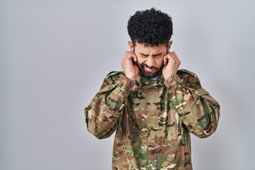 Arab man wearing camouflage army uniform covering ears with fingers with annoyed expression for the...