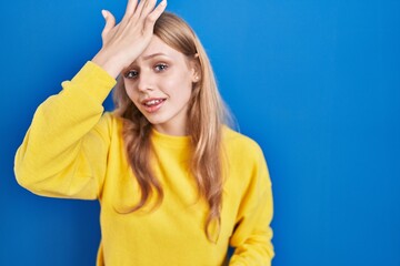 Young caucasian woman standing over blue background surprised with hand on head for mistake,...
