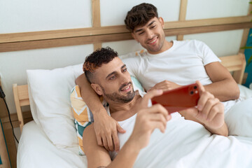 Fototapeta premium Two hispanic men couple lying on bed watching video on smartphone at bedroom