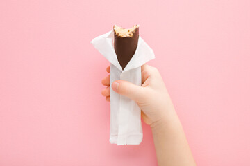 Little girl hand holding bitten dark brown chocolate bar with marzipan on light pink table...