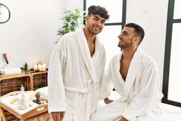 Two hispanic men couple wearing bathrobe sitting on massage table at beauty center