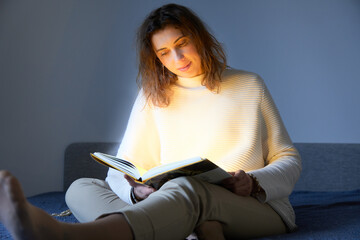 Beautiful woman illuminated by light from an open book.  Young pretty female reading book in cozy room.