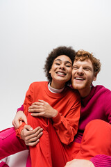 Obraz premium positive redhead man in magenta color sweatshirt hugging cheerful african american woman on grey.