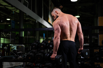 Fototapeta na wymiar A brutal muscular bearded man stands with his back and holds a dumbbell in the gym. Active lifestyle, sports