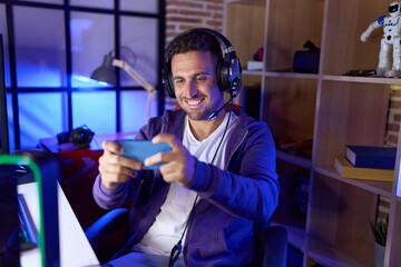 Young hispanic man streamer playing video game using smartphone at gaming room