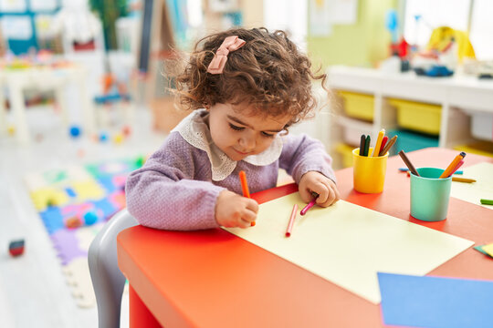 Adorable hispanic girl preschool student sitting on table drawing on paper at kindergarten