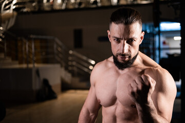 A handsome bearded male athlete with a muscular body shows his fist in the gym