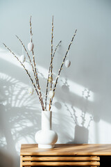 Easter home decor. Modern minimalist style interior with blooming branches of the pussy willow and hanging easter eggs in ceramic vase on a wooden console with sunlight and shadows on white gray wall. © okrasiuk