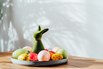 Easter home decor. Composition of Green bunny rabbit figurine and colored easter chocolate eggs on the plate on the wooden console with sunlight and shadows on white wall. Selective focus. Copy space.